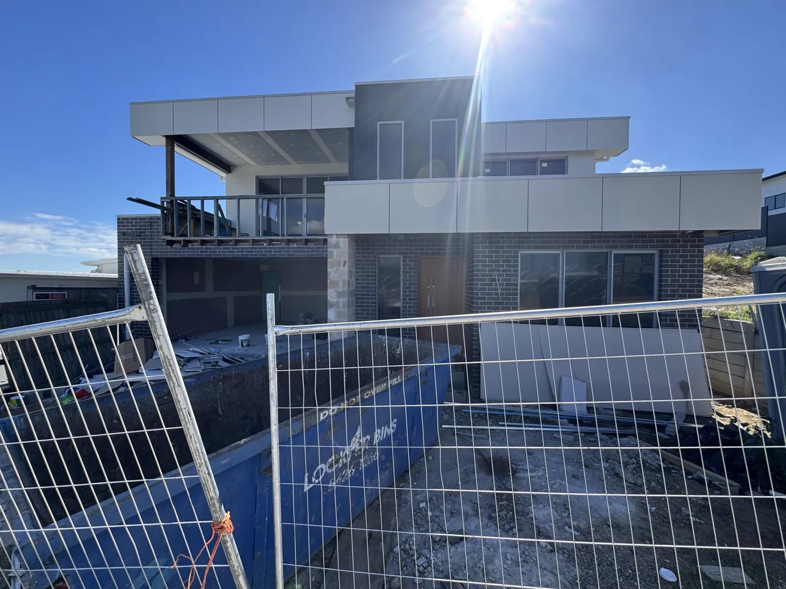 Construction progress photo of the 24 Delphinus Vista home taken head-on from the footpath directly in front of the house, showing the full width of the two-storey front elevation with the two upper-level balconies (small matte-black-framed glass balustrade balcony on the left, wider solid-parapet balcony on the right), central ebony vertical shiplap strip flanked by off-white Maxrix panels, symmetric matte-black-framed ground-floor sliding-glass units flanking the Oak pivot front door, temporary steel-mesh site fence and blue construction skip bin in the foreground.
