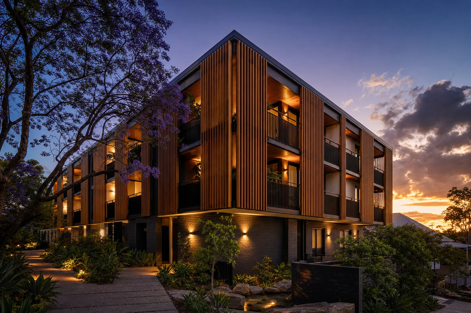 Small three-storey boutique apartment building at dusk with charcoal brick base, spotted-gum timber batten balcony screens, black steel balustrades, jacaranda overhead and warm interior lighting — indicative render of a Dream City Homes multi-residential project.