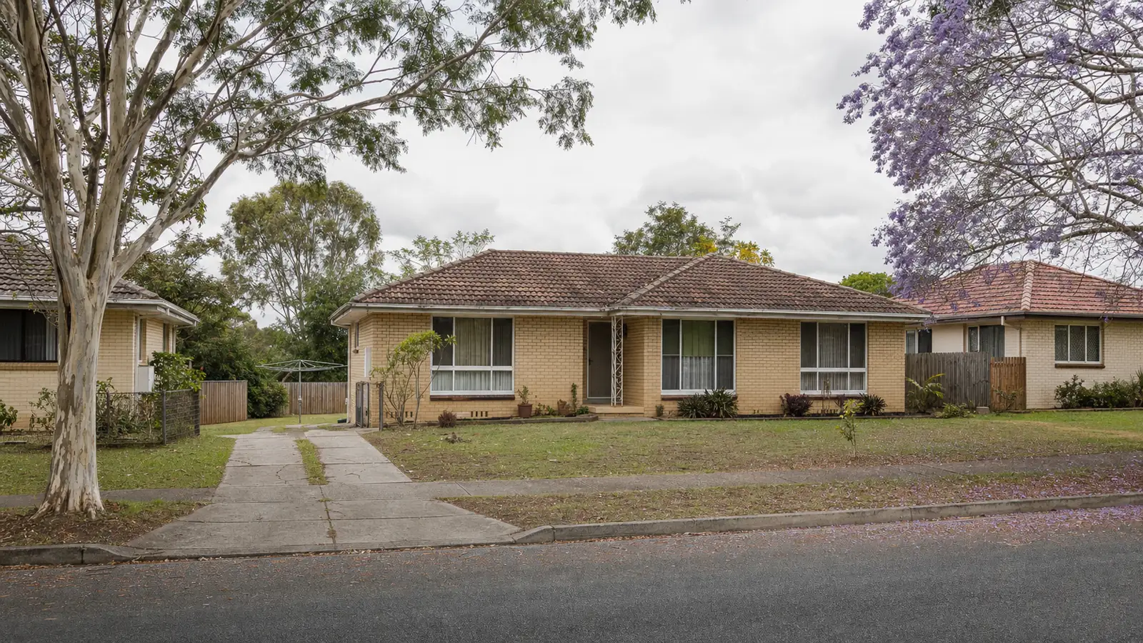 Existing 1970s brick-and-tile single-storey home on an established leafy Brisbane street before knock-down rebuild, weathered terracotta tile roof and pale brown brick.